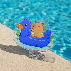 Puede incluir: Un portatarjetas de acrílico transparente con un capibara de dibujos animados con un collar de flores, flotando en un flotador de piscina con forma de pato azul. El portatarjetas tiene un clip de metal plateado y una correa de plástico transparente.
