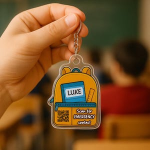Könnte beinhalten: Ein gelber, rucksackförmiger Schlüsselanhänger mit einem blauen Notizbuch mit der Aufschrift "LUKE" und einem Lineal. Der Schlüsselanhänger enthält einen QR-Code und den Text "Scan for EMERGENCY contact". Der Schlüsselanhänger wird von einer Person gehalten.