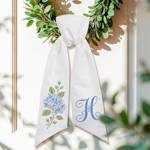 May include: White sash with blue embroidered hydrangea and the letter "H". The sash is hanging on a white door with a green wreath in the background. The hydrangea has blue petals and green leaves.