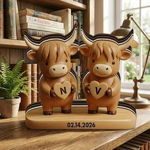 May include: Two wooden Highland cow figurines with heart-shaped cutouts, each holding a letter. The figurines stand on a wooden base with the date "02.14.2026" engraved. The cows have large horns with black and gold stripes.