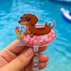 Pode incluir: Um porta-crachá transparente com um dachshund de desenho animado usando óculos de sol e flutuando em uma boia floral rosa. O cachorro está ao lado de um coquetel com uma fatia de laranja e um pequeno guarda-chuva. O porta-crachá tem uma alça de plástico transparente e um clipe de metal.