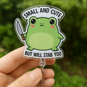 May include: A badge reel with a cartoon frog holding a knife. The frog is light green with pink cheeks and large black eyes. Text above reads "SMALL AND CUTE", and below reads "BUT WILL STAB YOU".