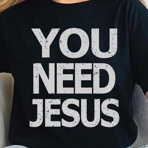 Könnte beinhalten: Schwarzes T-Shirt mit dem weißen Text "YOU NEED JESUS". Der Text hat ein abgenutztes, strukturiertes Aussehen. Das Shirt ist ein lässiger Stil, geeignet für den Alltag.