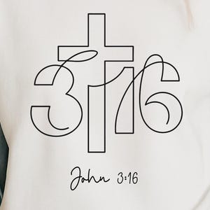Puede incluir: Sudadera blanca con un diseño de arte lineal negro que presenta una cruz, los números 3:16 y el texto "John 3:16". La sudadera está hecha de un material suave.