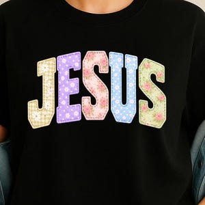 Könnte beinhalten: Schwarzes T-Shirt mit dem Wort "JESUS" in bunten Buchstaben im Patchwork-Stil. Die Buchstaben sind in verschiedenen Mustern gehalten, darunter Blumen, Vichy-Karo und Tupfen, mit einem genähten Rand. Das Shirt wird mit einer Jeansjacke getragen.