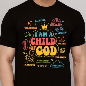 Könnte beinhalten: Schwarzes T-Shirt mit dem farbenfrohen Schriftzug "I AM A CHILD OF GOD". Das Design enthält auch Wörter wie "Geliebt", "Einzigartig", "Dankbar" und "Kostbar" in einem verspielten Stil.