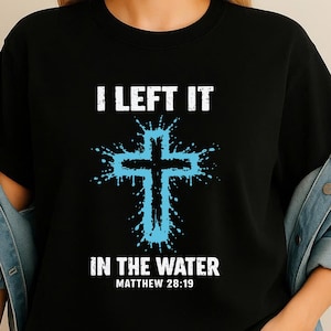 Puede incluir: Camiseta negra con el texto blanco "I LEFT IT IN THE WATER" y "MATTHEW 28:19". Un diseño de cruz azul con efecto de salpicadura está en el centro. La camiseta es de cuello redondo.