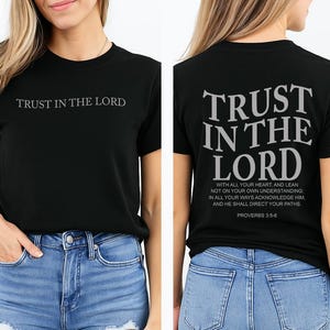 Può includere: T-shirt nero con la scritta "TRUST IN THE LORD" sul davanti e sul retro. Il retro della maglietta presenta lo stesso testo in un carattere più grande, insieme a un versetto biblico. La maglietta è abbinata a jeans blu.