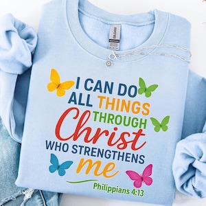 Könnte beinhalten: Hellblaues Sweatshirt mit dem Text "I CAN DO ALL THINGS THROUGH Christ WHO STRENGTHENS me" und bunten Schmetterlingen. Der Text "Philippians 4:13" ist ebenfalls auf dem Shirt aufgedruckt. Eine silberne Halskette liegt obenauf.