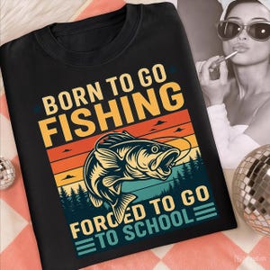 May include: Black crew neck t-shirt with a graphic design. The design features a fish jumping out of the water, with text that reads "BORN TO GO FISHING" above and "FORCED TO GO TO SCHOOL" below. The background has orange, yellow, and blue stripes.