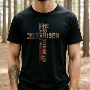 Könnte beinhalten: Schwarzes T-Shirt mit einem Holz-Kreuz-Design und dem Text "HE IS RISEN" und "MATTHEW 28:6". Das Kreuz enthält Elemente der amerikanischen Flagge, einschließlich Sterne und Streifen. Das T-Shirt hat einen Rundhalsausschnitt.