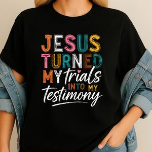 Könnte beinhalten: Schwarzes T-Shirt mit dem Text "JESUS TURNED MY TRIALS INTO MY TESTIMONY" in bunten Blockbuchstaben. Das Design enthält ein kleines rosa Herz. Das Shirt hat einen Rundhalsausschnitt und kurze Ärmel.