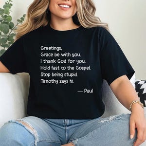 Könnte beinhalten: Schwarzes T-Shirt mit weißem Text. Der Text lautet: „Grüße, Gnade sei mit euch. Ich danke Gott für euch. Haltet am Evangelium fest. Hört auf, dumm zu sein. Timotheus sagt hallo. - Paul“. Ein lässiges Kleidungsstück.