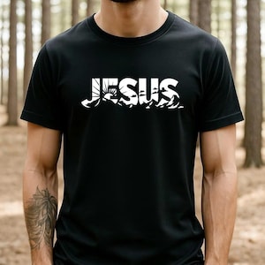 Könnte beinhalten: Schwarzes T-Shirt mit dem Wort "JESUS" in Weiß. Das Wort steht über einer weißen Grafik von Bergen, Sonne und Wellen. Das T-Shirt hat kurze Ärmel.
