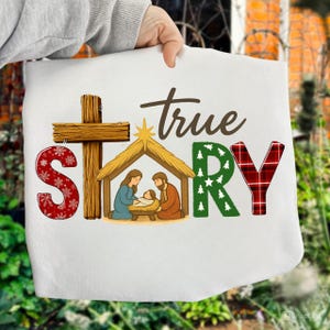 May include: White tote bag featuring a Nativity scene with the text "true story." The letter "S" is red with white snowflakes, "R" is green with white stars, and "Y" is red plaid. A wooden cross and stable are also depicted.
