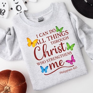 Könnte beinhalten: Ein hellgraues Sweatshirt mit dem Text "I CAN DO ALL THINGS THROUGH Christ WHO STRENGTHENS me" und bunten Schmetterlingsgrafiken. Der Text ist bordeauxrot und die Schmetterlinge sind gelb, grün, blau und pink. Das Sweatshirt ist von der Marke Gildan.