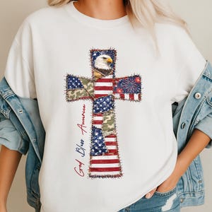Könnte beinhalten: Weißes Sweatshirt mit Rundhalsausschnitt und einem Kreuzdesign mit patriotischen Elementen. Das Kreuz besteht aus Mustern der amerikanischen Flagge, Tarnfarben und einem Weißkopfseeadler. Die Worte "God Bless America" sind in roter und blauer Schrift geschrieben.