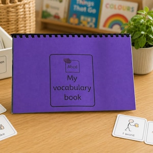 May include: A purple spiral-bound vocabulary book with a black rectangular border and the text 'My vocabulary book' and 'Abcd' on the cover, accompanied by several white square communication cards featuring stick figure illustrations and text.