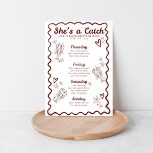 以下が含まれることがあります： ケープコッドでの独身女性向け週末の白い印刷された旅程で、赤い装飾的なボーダーが付いています。「She's a Catch」と書かれており、ロブスターとハートのイラスト付きのスケジュールが含まれています。