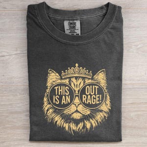 May include: Dark gray t-shirt with a gold illustration of a cat wearing glasses and a crown. The glasses have the text "THIS IS AN OUT RAGE!". The shirt is folded on a light wood surface.
