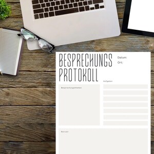 May include: A wooden desk workspace with a laptop, notepad and pen, glasses, a tablet, and a coffee cup. A document with the text "BESPRECHUNGS PROTOKOLL" is in the center.