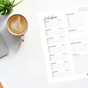 Könnte beinhalten: Ein weißer Wochenplaner für 2025 mit Abschnitten für jeden Tag, Prioritäten und Notizen. Ein Laptop, eine Kaffeetasse mit Latte Art und eine kleine Pflanze liegen auf einer weißen Oberfläche.