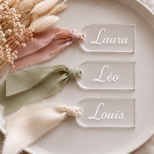 Peut inclure: Cartes de place en acrylique transparent avec les noms "Laura", "Léo" et "Louis" en écriture élégante. Chaque carte est attachée avec un ruban de couleur différente : rose poudré, vert sauge et crème. Les cartes sont disposées sur une assiette blanche avec des fleurs séchées.