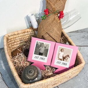 May include: A square woven basket containing two pink foam beverage insulators with black and white vintage photos and the text EST. 1982 and EST. 1988. Also included are a burlap-wrapped bottle, a small gray lidded jar, and a white rolled paper.