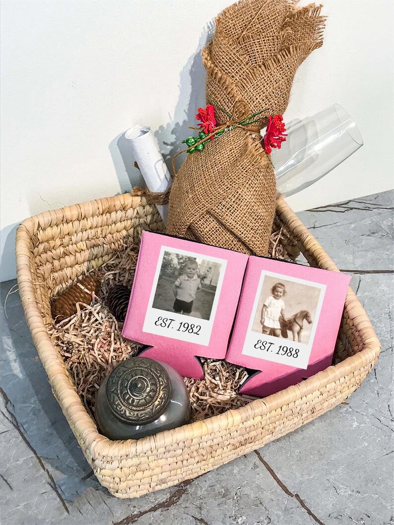 May include: A square woven basket containing two pink foam beverage insulators with black and white vintage photos and the text EST. 1982 and EST. 1988. Also included are a burlap-wrapped bottle, a small gray lidded jar, and a white rolled paper.