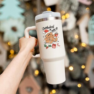 May include: A large, off-white travel mug with a white handle and lid. The mug features a Christmas-themed graphic of a gingerbread man wearing a Santa hat, holding a cup of coffee, and the text "Overbaked & over it."