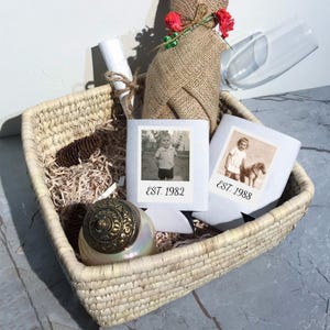 May include: A woven basket containing two white beverage insulators with vintage black and white photographs and the text EST. 1982 and EST. 1988. Also included are a burlap-wrapped bottle with red floral accents, a glass flute, and a round candle.