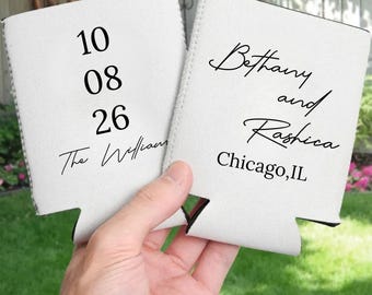 May include: Two white can coolers with black text. One cooler displays the date "10 08 26" and "The Williams". The other cooler reads "Bethany and Rashica" and "Chicago, IL". The coolers are held in a hand.
