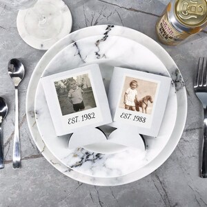 May include: Two white square beverage insulators featuring black and white vintage photographs of children. The left insulator displays the text EST. 1982, while the right insulator displays the text EST. 1988. Both items are positioned on a white marble plate.