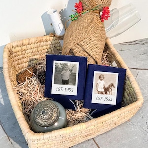 May include: Two navy blue foam can coolers featuring white square photo prints of children and the text EST. 1982 and EST. 1988. These items are nestled in a woven straw basket alongside a burlap-wrapped bottle, a small gray jar, and a glass.