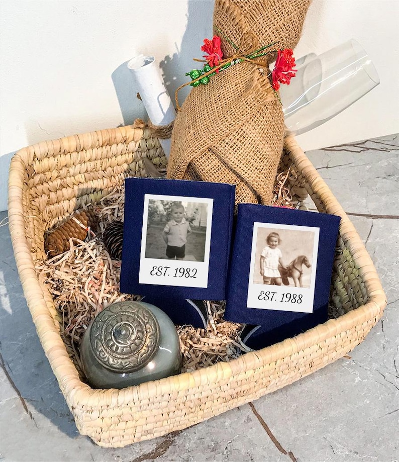 May include: Two navy blue foam can coolers featuring white square photo prints of children and the text EST. 1982 and EST. 1988. These items are nestled in a woven straw basket alongside a burlap-wrapped bottle, a small gray jar, and a glass.