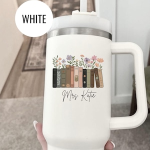 May include: White insulated tumbler with a handle and a floral and book design. The design includes a row of books with colorful flowers on top. The text "Mrs. Katie" is written in a script font.