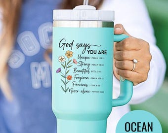 May include: A teal tumbler with a white lid and handle. The tumbler features the text "God says YOU ARE" with the words "Unique, Strong, Beautiful, Forgiven, Precious, Never alone" and corresponding Bible verses. A floral design accents the text.