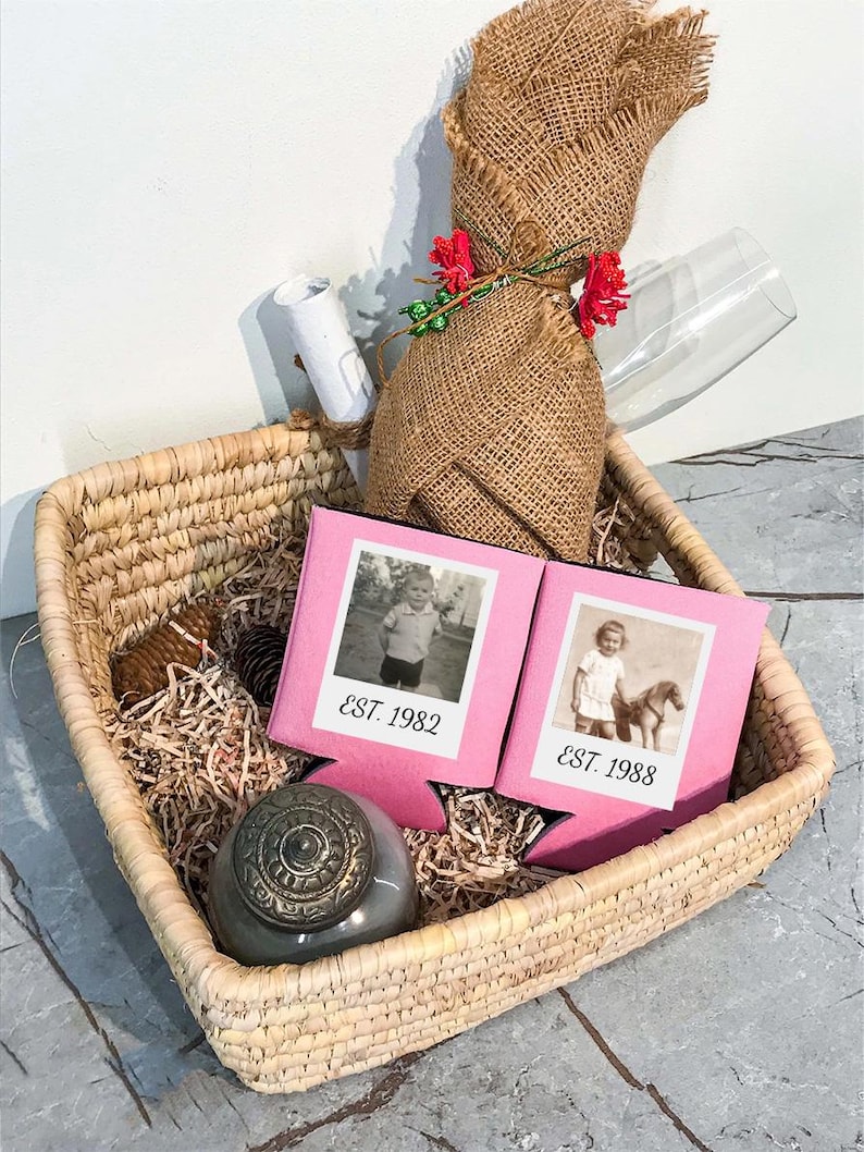 May include: A square woven basket containing two pink foam beverage insulators with black and white vintage photos and the text EST. 1982 and EST. 1988. Also included are a burlap-wrapped bottle, a small gray lidded jar, and a white rolled paper.