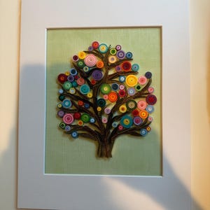 Può includere: Opera d'arte incorniciata in quilling di carta raffigurante un albero con tronco e rami marroni. Il fogliame dell'albero è composto da cerchi di carta arrotolata colorati in varie dimensioni e tonalità, su sfondo verde chiaro.