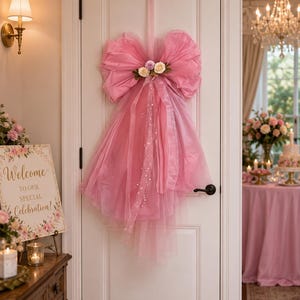 Pode incluir: Um grande laço decorativo de tule rosa pendurado numa porta branca. O laço apresenta tecido transparente em camadas, fitas longas e fluidas, e um grupo de pequenas rosas artificiais em tons de creme, rosa pálido e lavanda no centro.