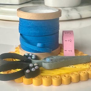 Könnte beinhalten: Eine handwerkliche Anordnung mit einer blauen Bandspule, einem rosa Maßband und einer schwarzen Schere, die auf einer gelben Papierfiligranbasis angeordnet sind. Die Szene deutet auf ein handgemachtes Projekt oder eine Bastelaktivität hin.