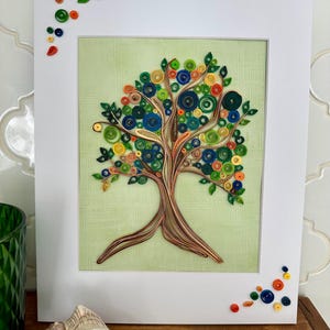 Könnte beinhalten: Gerahmtes Papier-Quilling-Kunstwerk mit einem Baumdesign. Der Baum hat einen braunen Stamm und Äste mit bunten, kreisförmigen Blättern in Grün, Blau, Orange und Gelb. Das Kunstwerk ist vor einem hellgrünen Hintergrund und in Weiß gerahmt.