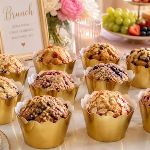 Pode incluir: Uma coleção de doze muffins em formas de papel dourado recortadas, com várias coberturas, incluindo mirtilos, framboesas e amêndoas laminadas, dispostos ao lado de uma placa com os dizeres Brunch Good Food Great Company Beautiful Day.