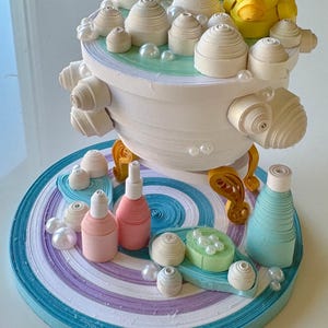 Puede incluir: Una caprichosa obra de arte de quilling de papel que representa una escena de baño. La bañera blanca, con patas doradas, está llena de burbujas de papel y un pato amarillo. La base es un remolino de azul, morado y blanco, con botellas y jabón de papel.