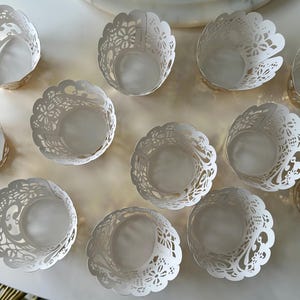 Puede incluir: Una colección de envoltorios de papel blancos para cupcakes, con diseños intrincados. Cada envoltorio presenta un delicado patrón de encaje floral y un borde festoneado. Los envoltorios están dispuestos sobre una superficie blanca, creando una exhibición visualmente atractiva.