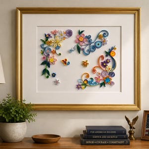 May include: A framed paper quilling art piece featuring intricate floral and swirling vine designs in shades of blue, pink, yellow, purple, and green. The artwork is displayed within a gold-toned frame against a white mat board background.