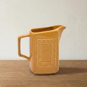 May include: Mustard-coloured ceramic jug with a rectangular handle and a spout. The jug features a recessed square design with a repeating geometric pattern. The jug is sitting on a wooden surface.
