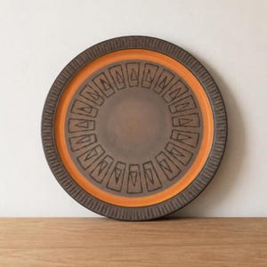Puede incluir: Un plato redondo de cerámica con un centro marrón y una banda naranja vibrante. El plato presenta un borde marrón oscuro texturizado y un patrón geométrico repetitivo en negro. El plato se exhibe sobre una superficie de madera.