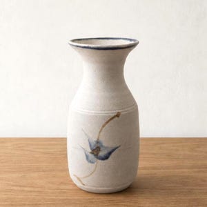 May include: A ceramic vase with a speckled off-white finish, featuring a flared rim with a thin blue border. The body of the vase is decorated with a hand-painted blue flower design on a slender brown stem, showcasing a textured, matte surface.
