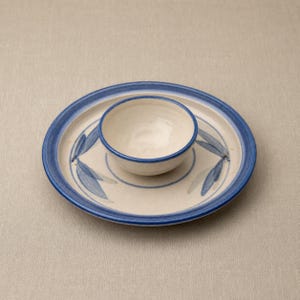 May include: A ceramic serving set comprising a small bowl on a larger plate. Both are off-white with blue detailing. The plate has a blue rim and leaf patterns. The bowl also has a blue rim. A simple, elegant design, suitable for serving.